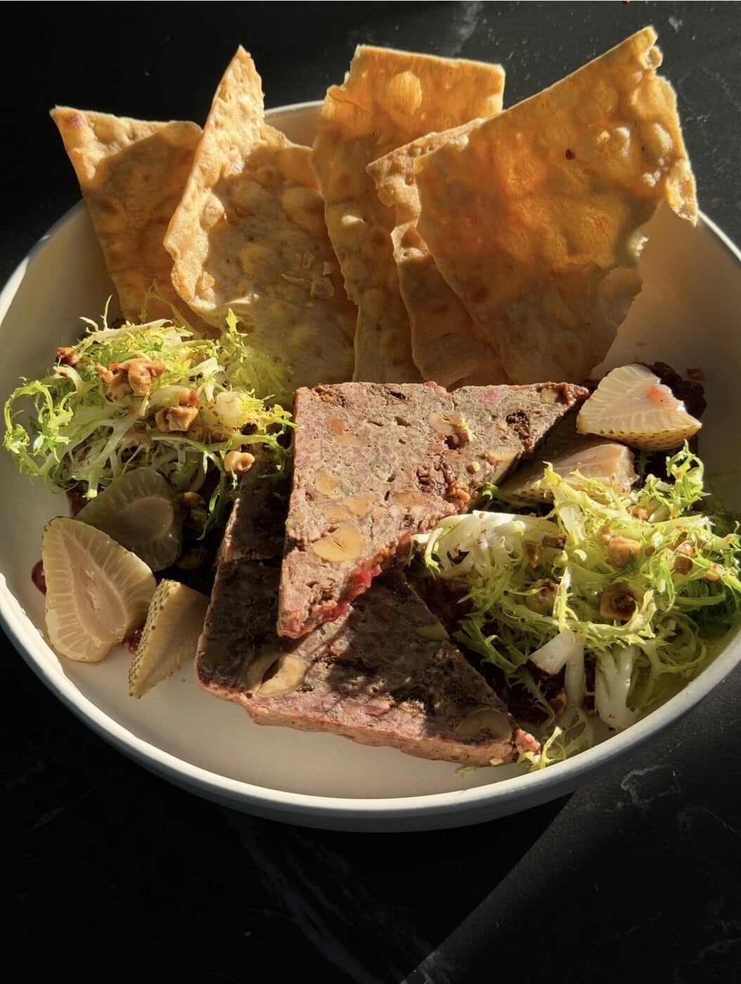 A White Bowl With Crisp Flatbread, Two Triangular Slices Of Pâté, Pickled Radishes, Frisée Lettuce, And Toasted Nuts, All Arranged On A Dark Surface In Sunlight.