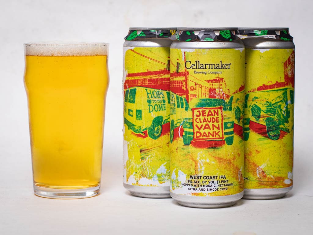 A Pint Glass Of Hazy Yellow Beer Sits Next To Three Cans Of Cellarmaker’s Jean Claude Van Dank West Coast Ipa, Featuring A Colorful, Abstract Label With Vans And Bold Yellow, Green, And Red Hues.