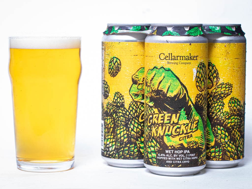 A Pint Glass Of Pale Beer Sits Next To Three Cans Of Green Knuckle Citra Ipa By Cellarmaker Brewing Co., Featuring A Green Fist With Hops On A Yellow Background.