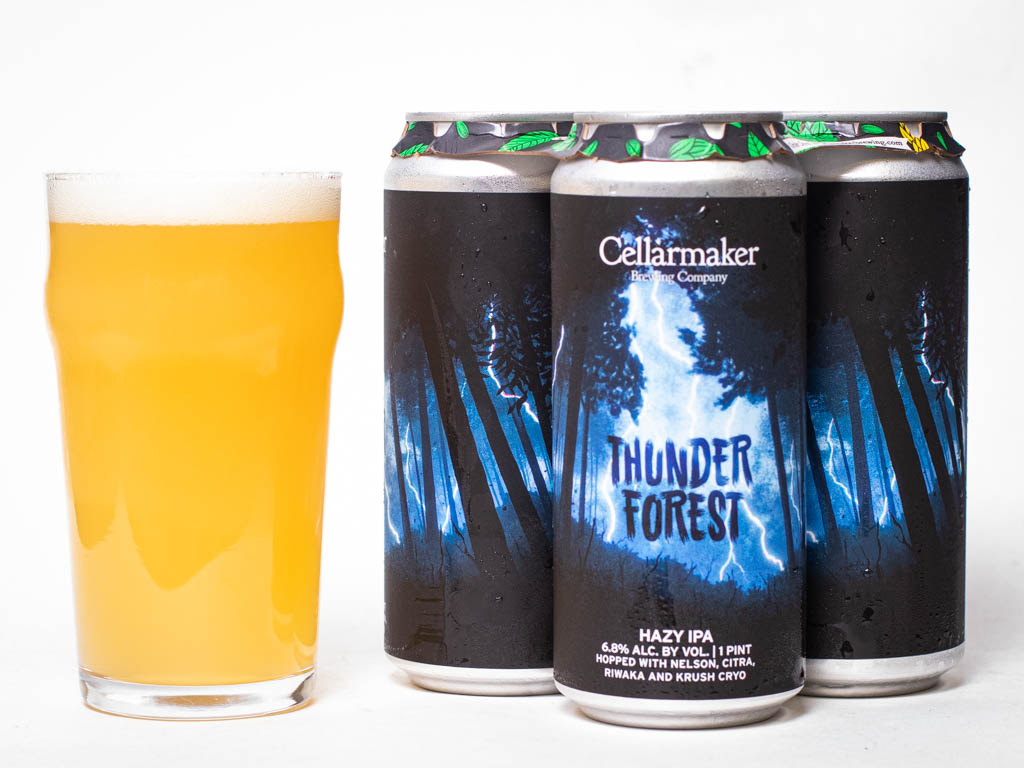 A Pint Glass Of Hazy Yellow Beer Sits Next To Three Cans Labeled “Thunder Forest” Hazy Ipa From Cellarmaker Brewing Company, With A Dark, Forest-Themed Design And Blue Lightning.