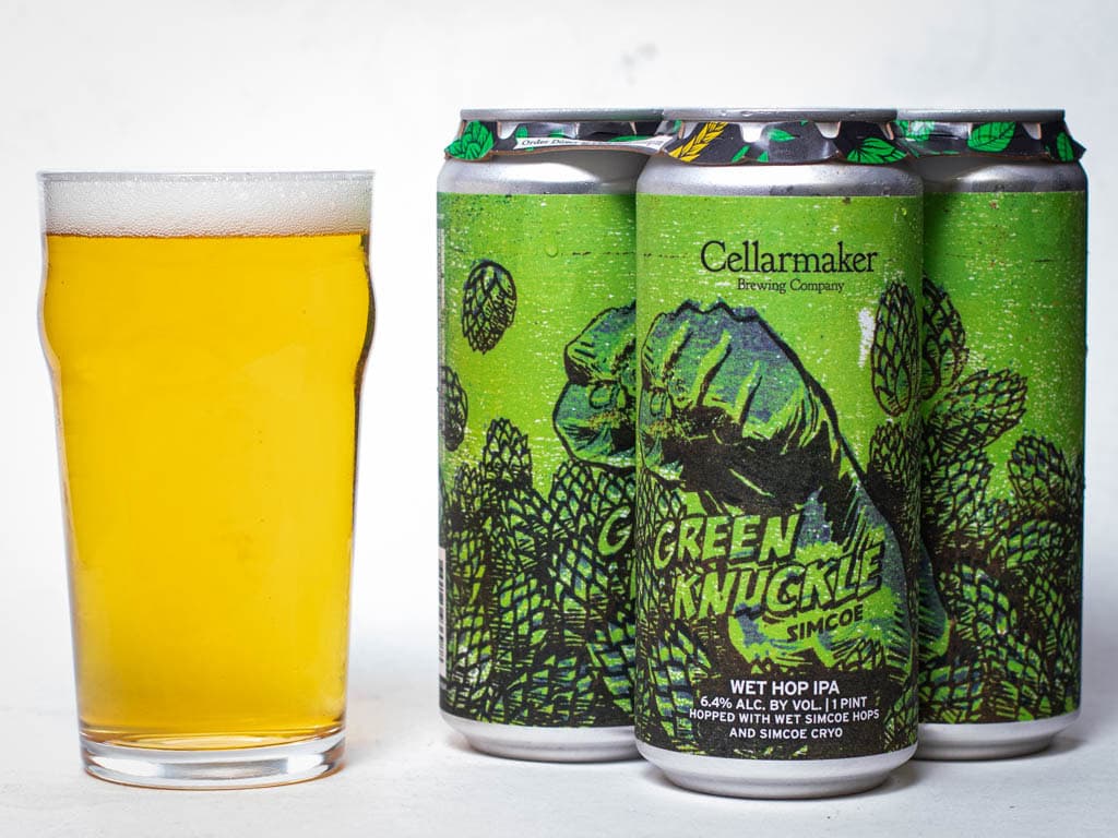 A Pint Glass Of Pale Beer Sits Next To Four Cans Of Cellarmaker’s Green Knuckle Simcoe Wet Hop Ipa, Featuring Green Hop Artwork And A Fist On The Labels.