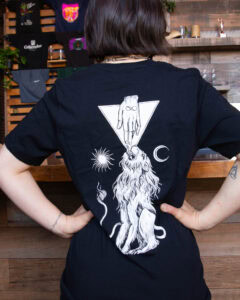 A person wearing a black Best Fiends Shirt featuring a back graphic of a hand reaching down from a triangle to touch the head of a lion, flanked by a sun and a crescent moon.