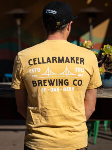 A person wearing a Classic Mustard Tee, a mustard-yellow t-shirt with a black graphic on the back featuring the text “Cellarmaker Brewing Co.” and an illustration of a bridge.