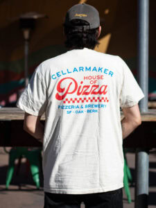 A person seen from the back wearing a cream-colored Double Dobis Shirt with “Cellarmaker House of Pizza” in red and blue lettering above a checkered pattern.