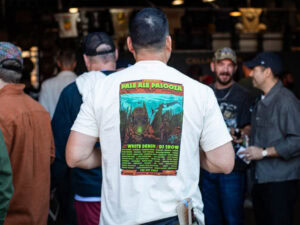 A person seen from the back wearing a white Pale Ale Palooza Shirt featuring a graphic of a deep-sea diver and a list of musical performers.
