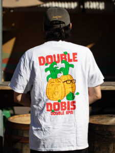 A person wearing a Double Dobis Shirt, which features a white t-shirt with a graphic of two pineapples with human faces, one wearing glasses, above the text “Double Dobis Double IPA.”