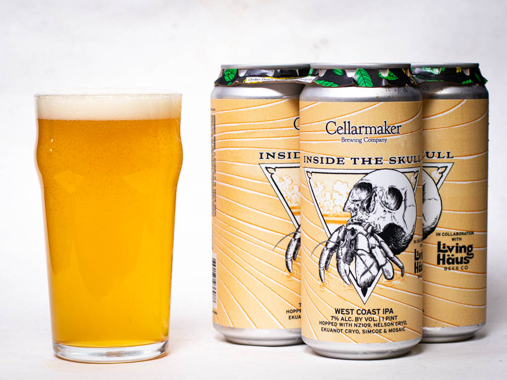 A Glass Of Golden Beer Next To A Four-Pack Of Cellarmaker Inside The Skull West Coast Ipa Cans.